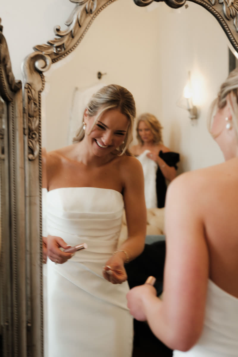 Bride laughing in a getting-ready room, her mother holding the dress behind her